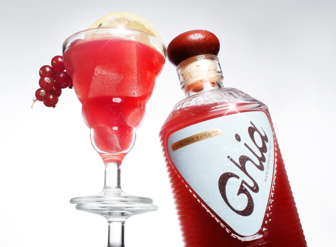 Frothy red Ghia cocktail garnished with red currants beside a Ghia apéritif bottle