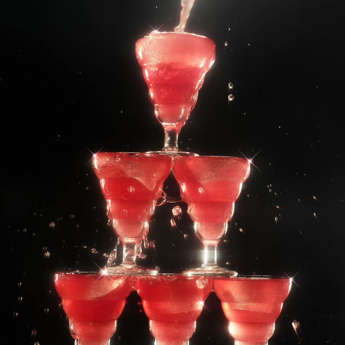 A tower of six glasses filled with deep red Ghia Le Fizz sparkling aperitif, with liquid splashing as it's poured into the top glass, creating a lively and sparkling effect.