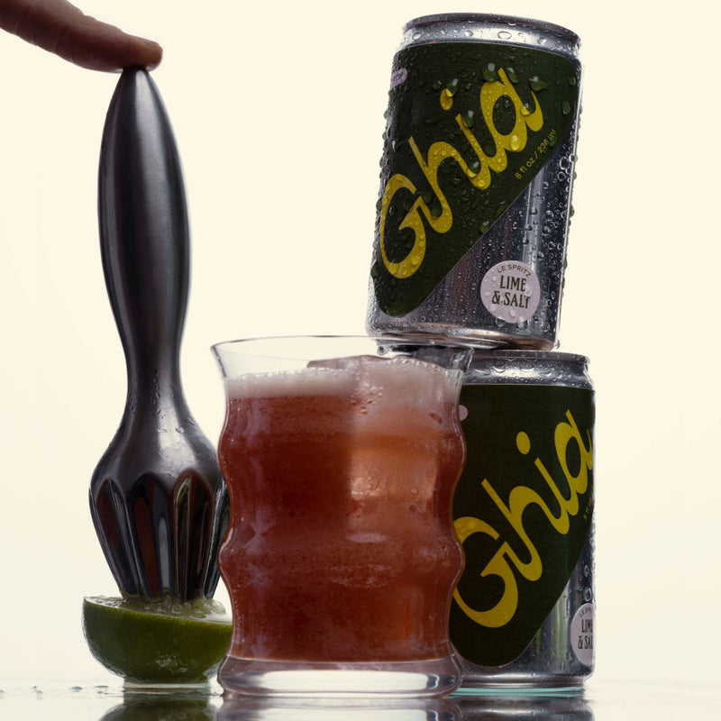 Two cans of Ghia Lime & Salt stacked together with condensation. A glass filled with the drink, and a hand squeezing lime with a juicer in the foreground.