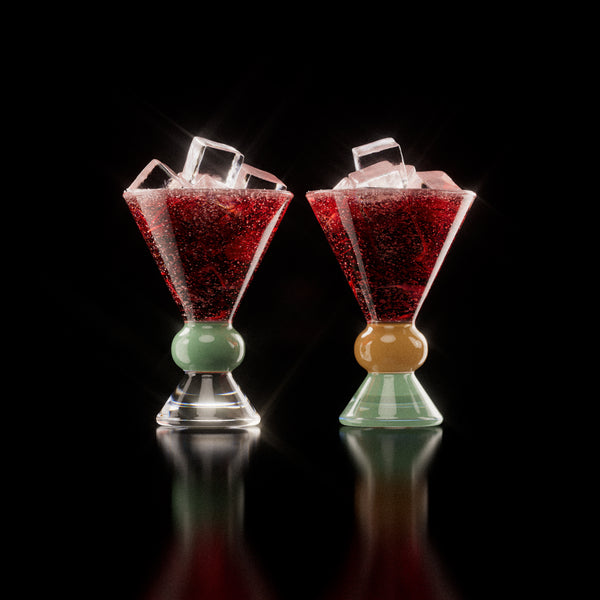 Two decorative glasses, each containing a red drink with ice cubes, highlighting the distinctive totem-inspired shape of the glasses against a light background with a clear, jade and sand base.