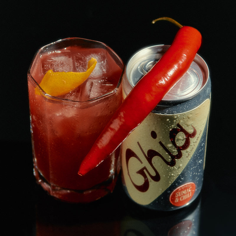 A can of Ghia Sumac & Chili non-alcoholic spritz with a red chili pepper resting on top. A glass of the drink with ice and a twist of orange peel is placed beside the can.