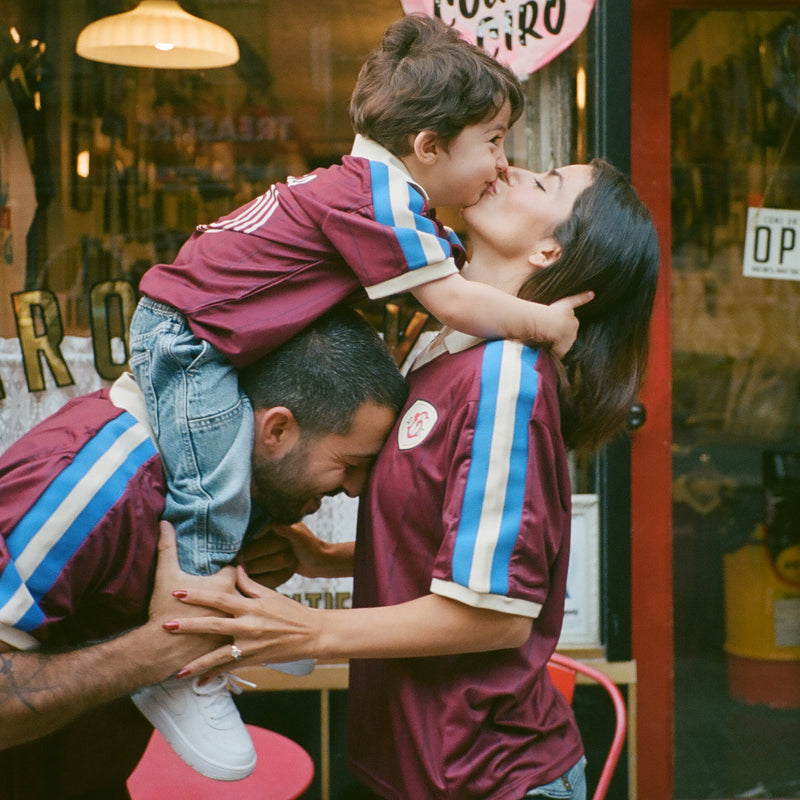 Family in maroon Ghia Jerseys, with a child on dad's shoulders leaning down to kiss mom
